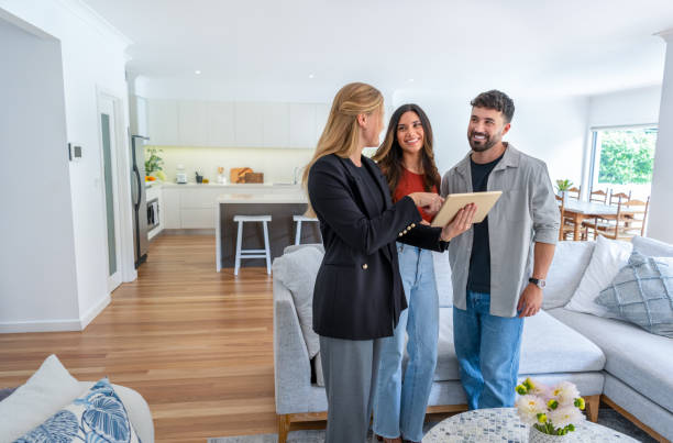 Real estate agent or architect showing a couple a new home. The agent is holding a digital tablet which everyone is looking at. The living room, kitchen and dining room can be seen in the background.