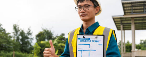 permit expeditor near me in los angeles : A male architect wearing a hard hat and safety vest checks his laptop while holding a clipboard with house plan papers. He reviews blueprints, construction documents,house structure details on-site.