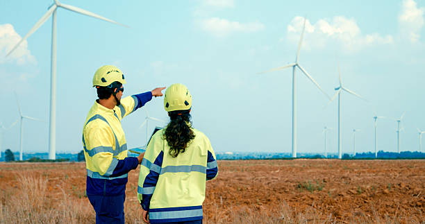 Renewable energy engineers are intently reviewing data on a laptop, contemplating the expansive turbine field before them. Green ecological power generation wind sustainable energy concept.