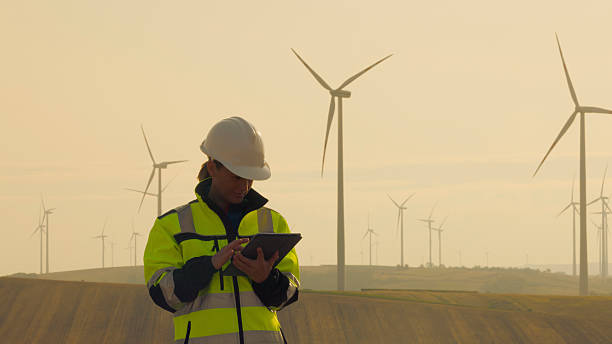 A mid adult Caucasian female technician in a high-visibility jacket and helmet uses a digital tablet at an outdoor wind power station during sunset.