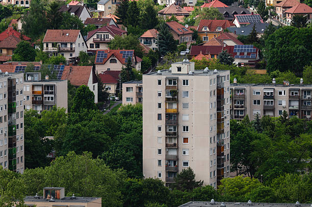 Four mid-rise apartment buildings in soft yellow and white tones stand in contrast to the lush green hillside behind them. Trees frame the scene naturally, blending urban residential life with the surrounding rural environment. The peaceful setting captures the edge of city expansion into nature.