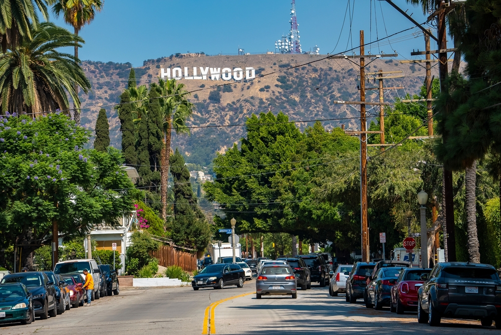 Los Angeles, USA. September 20, 2022. Vehicles moving on street surrounded with lush trees leading towards Mount Lee with Hollywood sign on sunny day with sky in background