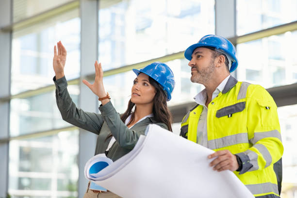 Young woman architect explaining blueprint to supervisor wearing safety vest at construction site. Mid adult contractor holding blueprint and understanding manager vision at construction site. Smiling engineer with hardhat on head talking to contractor while standing in building in construction process. benefits of hiring construction consultant