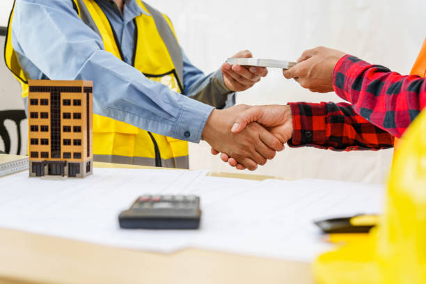 Two men, including a male Asian construction contractor engineer, discuss structural plans at a desk, engineering concrete columns and beams for a new house, paying dollar bills, and shaking hands.