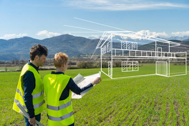 A couple are standing in a large field with hills and mountains in front of them as they hold up their house plans and try and imagine their new property, with a drawing of a house imposed onto the picture