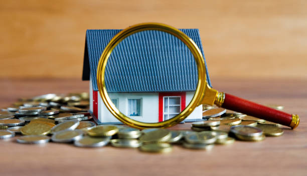 A house on coins, a magnifying glass highlighting details – a symbol of real estate appraisal, hidden costs, and potential defects that should be checked before buying