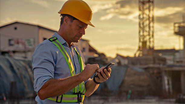A male construction worker wearing a yellow helmet and reflective vest checks his smartphone at an active construction site during sunset.