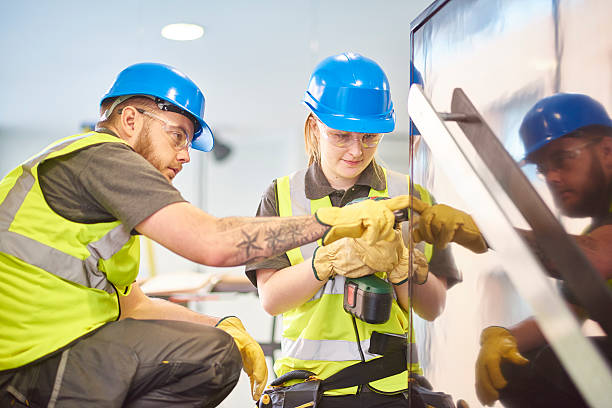 a teenage female apprentice carpenter is installing handrails in a modern office refurbishment. She is being supervised by her co-worker . She is using a drill to make guide holes for the hand rails . They are wearing safety workwear.