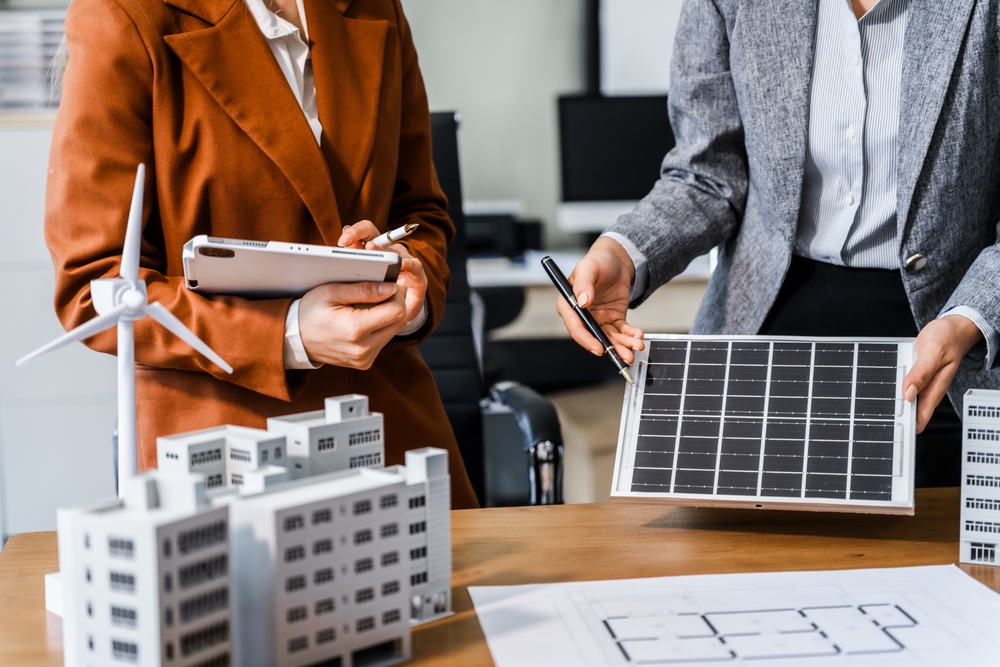 Two businesswomen sit at a desk in a meeting, discussing property development while building a model of city tower. real estate investments, zoning laws, sustainable energy solutions. net zero