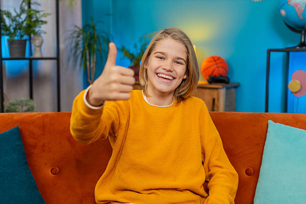 Like. Caucasian teenager boy gives thumbs up to camera while smiling at home. Sits comfortably on sofa, appearing satisfied with the good positive result, displaying feelings of approval, contentment