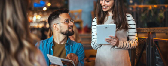 A smiling waitress takes an order from guests at a cozy bar, with one man holding a menu and engaging in lively banter dressed in casual attire.