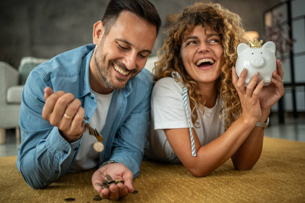 A happy young couple saving money for their next investment, purchase or vacation, they are lying on the living room floor and putting small coins in their small home safe