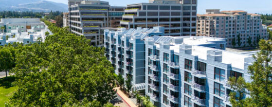 Aerial view of Cupertino's evolving cityscape showcasing a mix of modern architecture in California towns. Foreground features a sleek white residential complex with balconies and solar panels. Behind it, taller office buildings rise, their glass facades reflecting the California sun. Lush green trees line the streets, providing a natural contrast to the urban structures. The scene captures the essence of Silicon Valley's rapid growth and technological influence on urban planning.