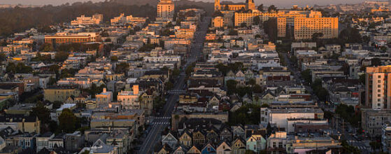 Aerial view of Mission District in San Francisco at sunrise with prominent church spires visible against pink morning sky. Straight road cuts through dense residential neighborhood with mix of Victorian houses and apartment buildings. Golden sunlight illuminates facades while Buena Vista Park creates dark silhouette in background. Historic architecture and urban planning of San Francisco visible from high angle view.