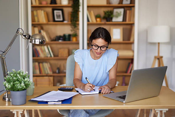Young woman working from her home office, taking notes while using her laptop