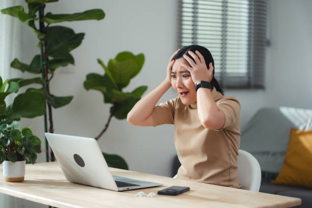 Asian woman sitting at home office thinking about problem with work, shock and feels concerned while looking at laptop.